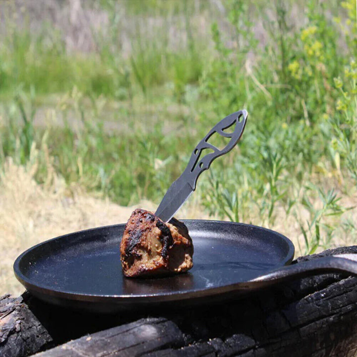 Black frying pan with a piece of toast and a knife on a log against a grassy background