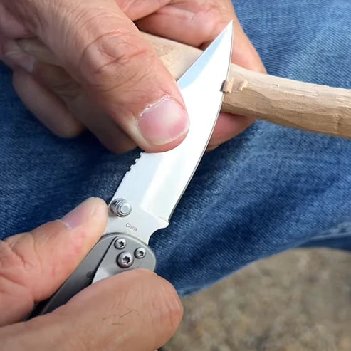 Person using a folding knife to cut a piece of wood against a blue jeans background
