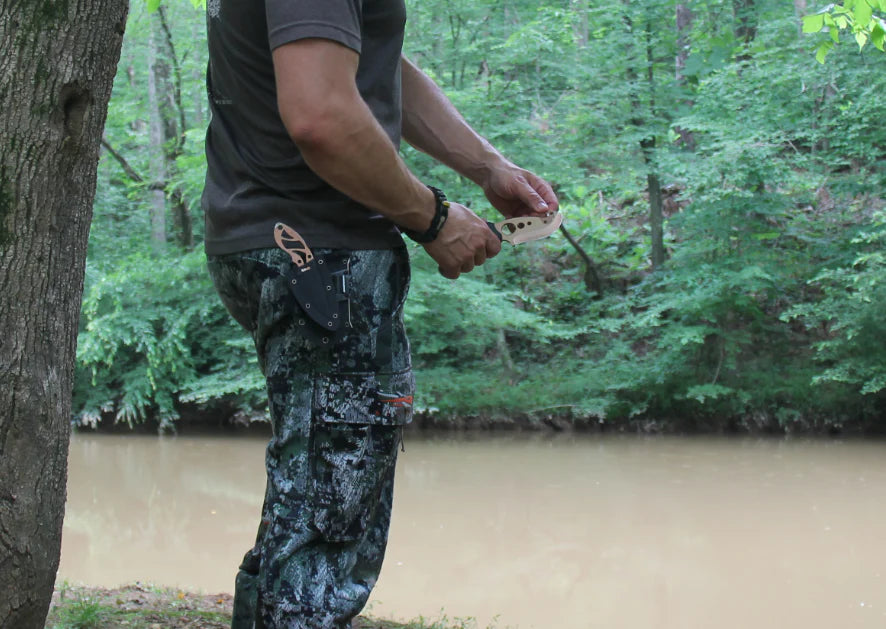 Person in camouflage pants and a dark shirt standing by a river with trees in the background